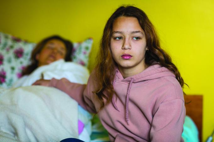 A young carer sits besides a bed where her mother is sleeping.