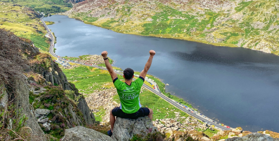 Man in Barnardo's jersey raising arms in victory on top of a mountain